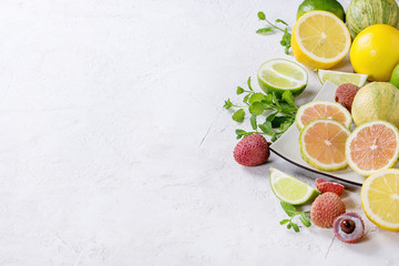Variety of whole and sliced citrus fruits pink tiger lemon, lemon, lime, mint and exotic lichee on white square plate over white concrete textured background. Copy space. Healthy eating