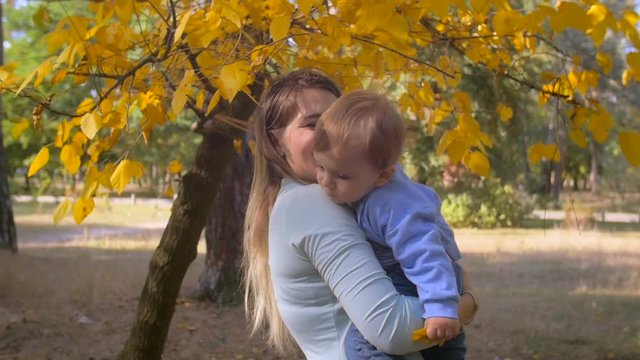 Slow motion steadicam shot of beautiful smiling mother cuddling her baby under tree at autumn park