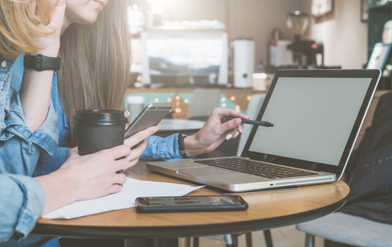 Two young business women sitting at table in cafe, drinking coffee and talking. First woman holding pen and smartphone.On table laptop and cup of coffee. Students learn online. Meeting of two friends.