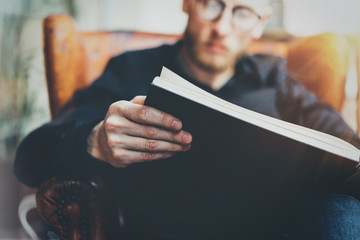 Closeup view of male hands holding black book.Young bearded man relaxing at home while sitting in vintage chair.Selective focus on hand,blurred background.Horizontal, film effect.