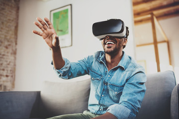 Concept of technology,gaming,entertainment and people.Happy bearded african man enjoying virtual reality glasses while relaxing on sofa at home.Blurred background.