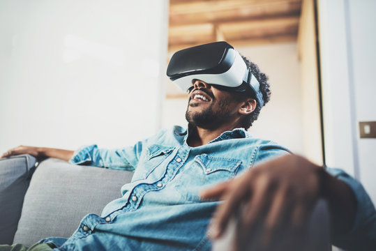Concept of technology,gaming,entertainment and people.Bearded african man enjoying virtual reality glasses headset or 3d spectacles while relaxing on sofa at modern home.Blurred background.