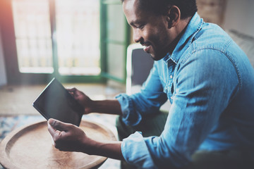 Closeup view of young bearded African man using tablet while sitting on sofa at home.Concept people working with mobile gadget.Blurred background, flares