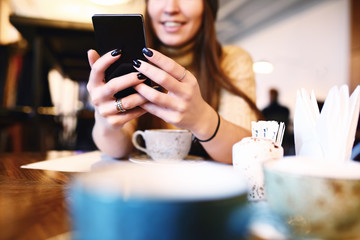 Woman typing text message on smart phone in a cafe. Cropped image of young woman sitting at a table with a coffee using mobile phone. Tonned. Selective focus.