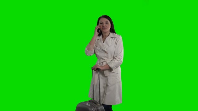 Woman with suitcase talking on the phone waving her hand saying hello against green screen