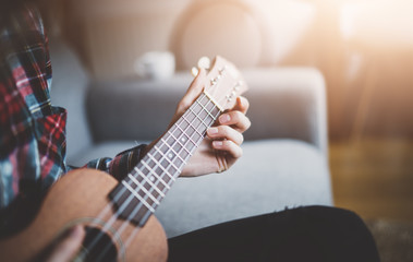 Fototapeta premium Young hipster girl playing ukulele guitar at sunny morning, close-up of female hands playing guitar, focus on the hands, flare light, film effects