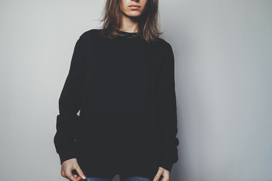 Young hipster girl wearing black oversize women’s hoodie with blank space for your logo or design, mock-up of black cotton sweatshirt, white wall in the background
