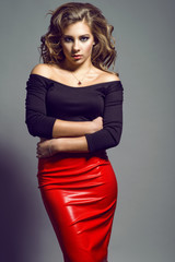 Portrait of young pretty model with long wavy hair wearing black top with naked shoulders and three quarter sleeve, red leather skirt and sterling silver necklace pendant embracing herself