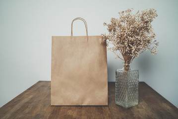 Mock-up of brown shopping bag on wooden table with glass vintage vase, blank craft package with handles with empty area for your design or logo