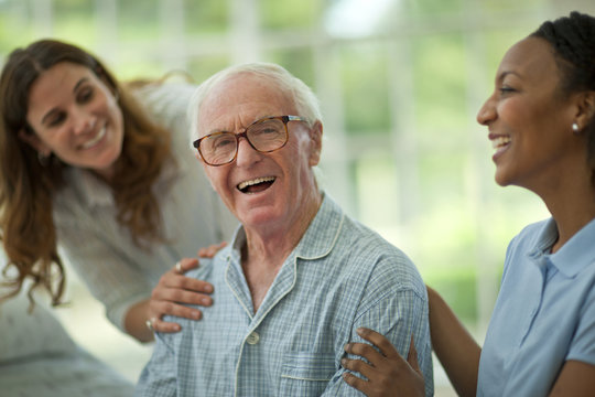 Portrait of a laughing senior man being comforted by a female nurse and his adult daughter while wearing pajamas.