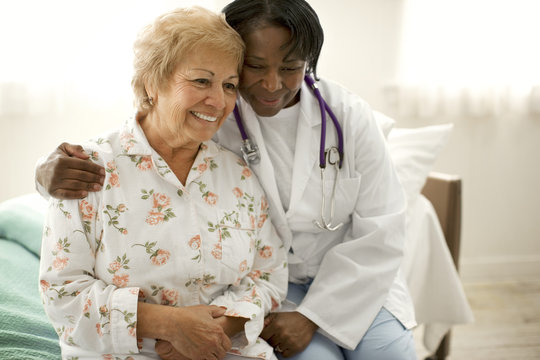 Smiling senior woman sitting next to a female doctor on the edge of a bed.
