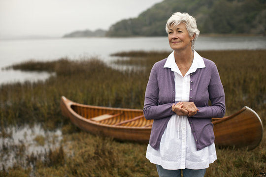 Mature woman looks away as she stands with her hands clasped posing for a portrait in front of a wooden canoe on a lakeshore.