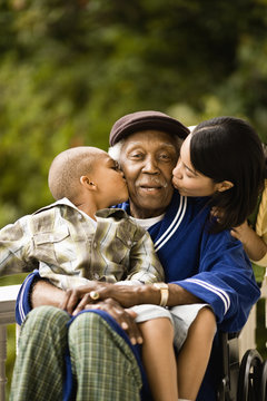 Senior man sitting in wheelchair is kissed on the cheek by his grandson sitting in his lap and on the other cheek by a young woman leaning over his shoulder as they pose for a portrait on a porch.