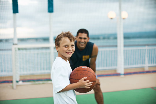 Boy holding a basketball and his basketball tutor smile in the background