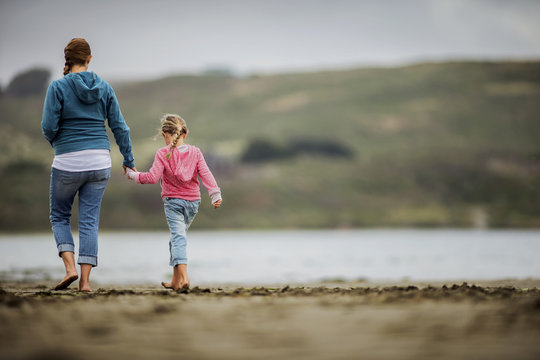Young blond girl walks hand-in-hand with her mother along the beach.
