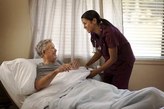 Smiling young nurse tucking in an elderly patient in a hospital bed.