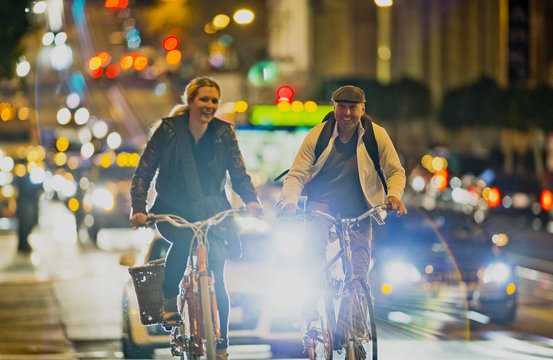 Middle-aged man and his pretty young girlfriend smiling as they cycle together along a busy city street at night.