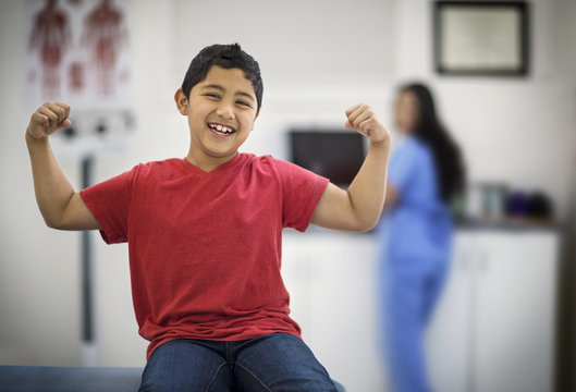 Portrait of a laughing young boy flexing his muscles while sitting inside a doctor's office.