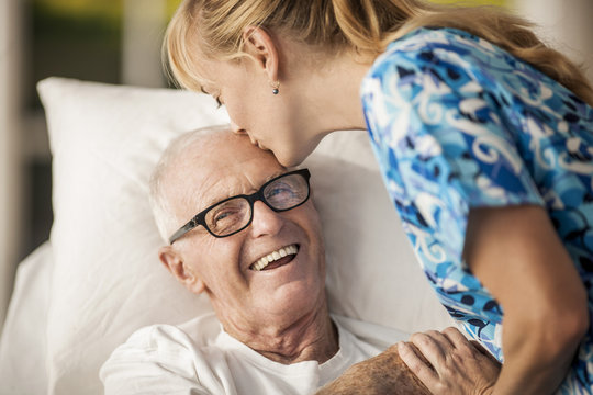Friendly young rest home nurse cheers up her elderly male patient with a kiss on his forehead.