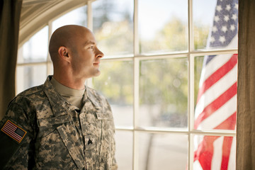 Thoughtful soldier looking through a window while standing inside his home.