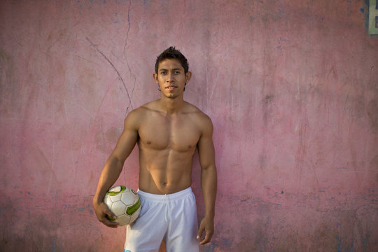 Portrait of a shirtless young man holding a soccer ball while standing against a wall.