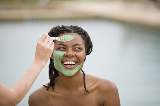 Young woman with bare shoulders smiling while having a mud face mask applied.