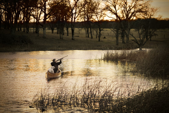 Man and his dog on a canoe with a gun along a river at sunset.