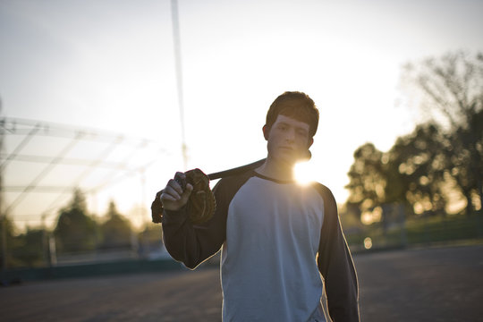Portrait of a teenage boy carrying a baseball mitt and baseball bat over his shoulder in the sunlight.