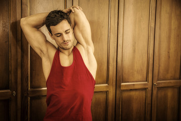 Fototapeta na wymiar Sexy handsome young man standing, wearing red tank-top in his bedroom against wooden wardrobe door
