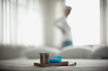 Two pill bottles,  a medication dispenser and two books sitting on a bed with a woman standing in front of a window in the background.