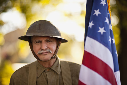 Mature male reenactor wearing an American World War steel combat helmet and uniform and holding an American flag poses for a portrait.