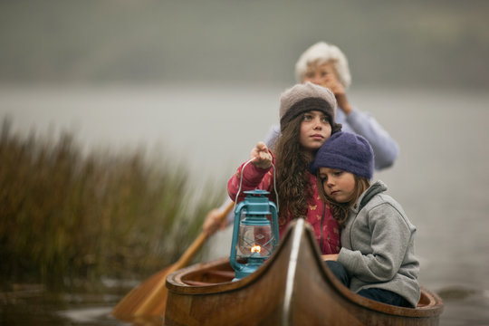 Solemn young girl holds a lit lantern and sits with her arm around her tired younger sister at the prow of a wooden canoe as a woman paddles them past reeds towards to lakeshore.
