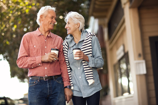 Smiling senior couple walk down the street drinking coffee and looking at each other.