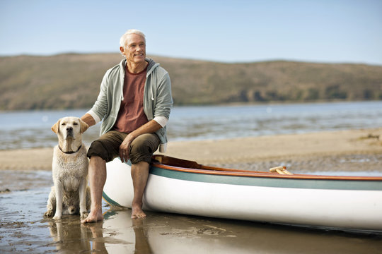 Happy senior man sitting on the edge of a canoe with his dog.