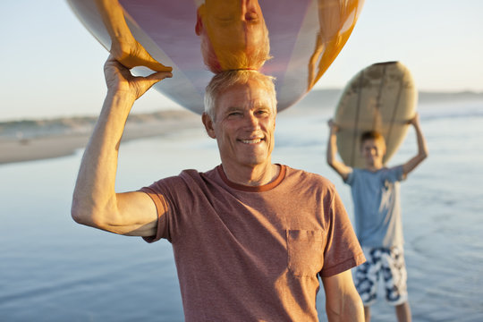 Portrait of a smiling senior man and grandson carrying surfboards on top of their heads.