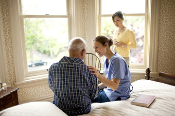 Unwell senior man receiving a visit from his nurse and his adult daughter.