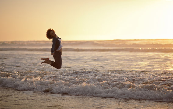 Portrait of a laughing woman leaping into the air while standing at the edge of the sea on a beach.