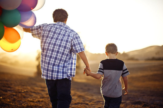 Two brothers walking holding hands, while one holds a large bunch of balloons.