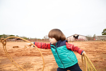 Handsome small toddler boy training making loope and throwing lasso in ranch. Red clay background,...
