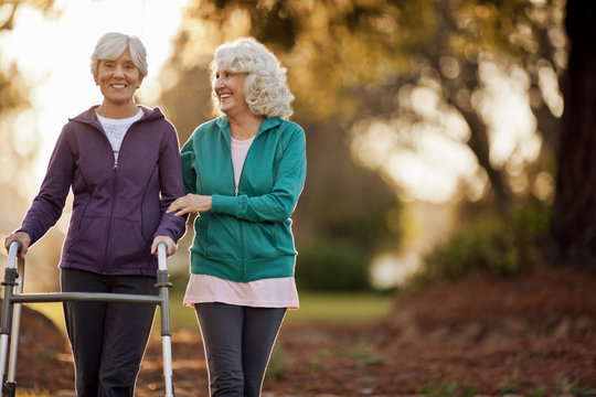 Portrait of a smiling senior woman being helped by her friend to walk with a walking aid through a park.