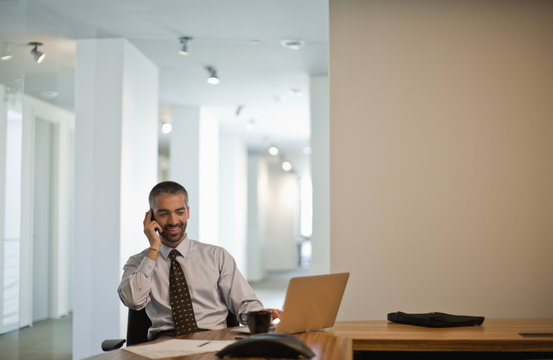 Businessman sits at desk talking on cellphone.