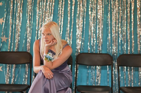 Teenage girl sitting on chair in evening gown, looking bored at prom.