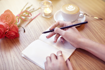 Woman hands close-up writing in notebook, using pencil, blank pages for layout. Cappuccino latte coffee with heart on top. Flowers on wooden table in modern cafe. St. Valentines celebration concept