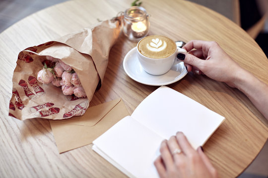 Woman hands close-up writing in notebook, using pencil, blank pages for layout. Cappuccino latte coffee with heart on top. Flowers on wooden table in modern cafe. St. Valentines celebration concept