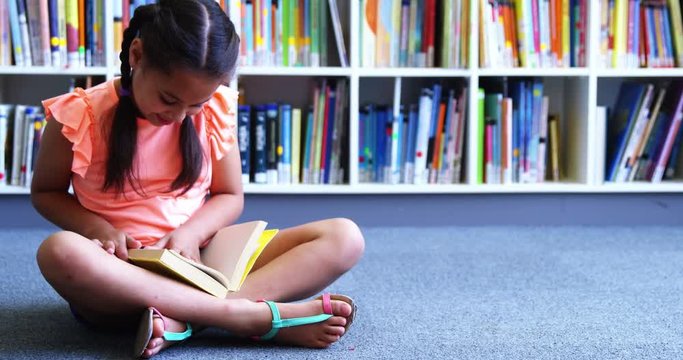 Slow motion of schoolgirl sitting on floor and reading a book in library at school 4k