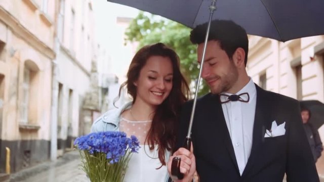 Few shots of young couple walking down the old citys street under umbrella, laughing and being happy together. Attractive young European girl in a white dress. Handsome young man in an elegant suit.