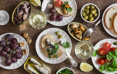 Seafood snacks table - canned sardines, mussels, octopus, grape, olives, tomato and two glasses white wine on wooden table, top view. Flat lay