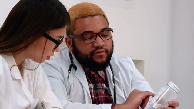 Afroamerican male doctor holding jar of pills while nurse making notes