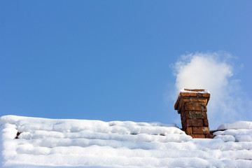 smoke out of a brick chimney on a snowy rooftop on the background of blue sky