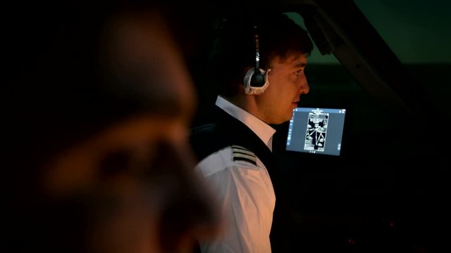 Close shot of two professional pilots at the cockpit of modern passenger airliner. Aircraft taxiing at the runway. Captain talks by radio with air traffic controller at tower. Shot at night or early